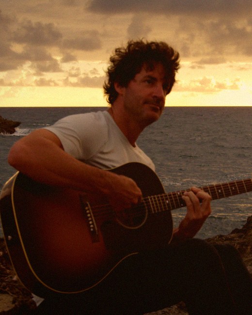 HINZE seated with an acoustic guitar as the sun sets over the ocean